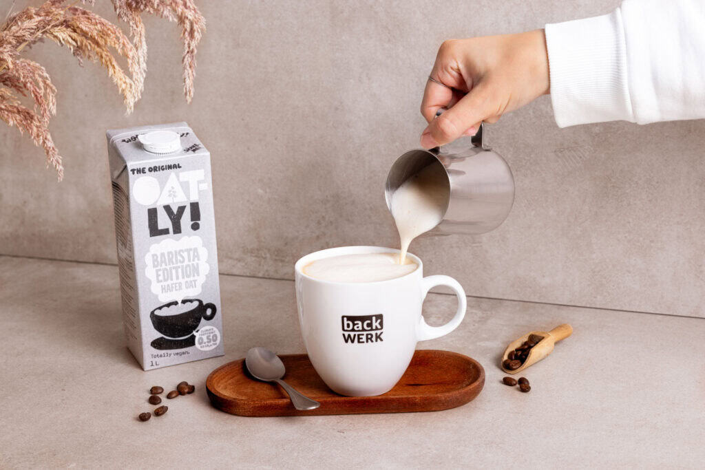 Backwerk tasse wird mit augeschäumter milch befüllt, daneben ein karton oatly barista